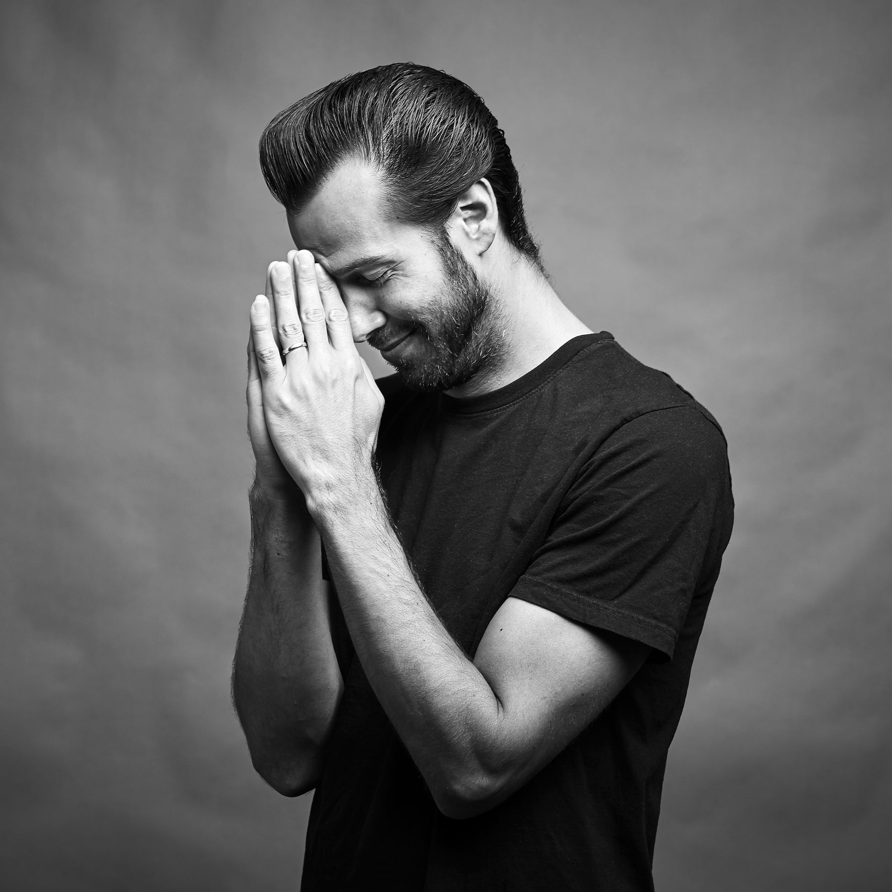Black and white portrait of a man in a black t-shirt with slicked-back hair and beard, smiling with eyes closed, hands pressed together in front of face in a reflective or thankful gesture, set against a neutral background.